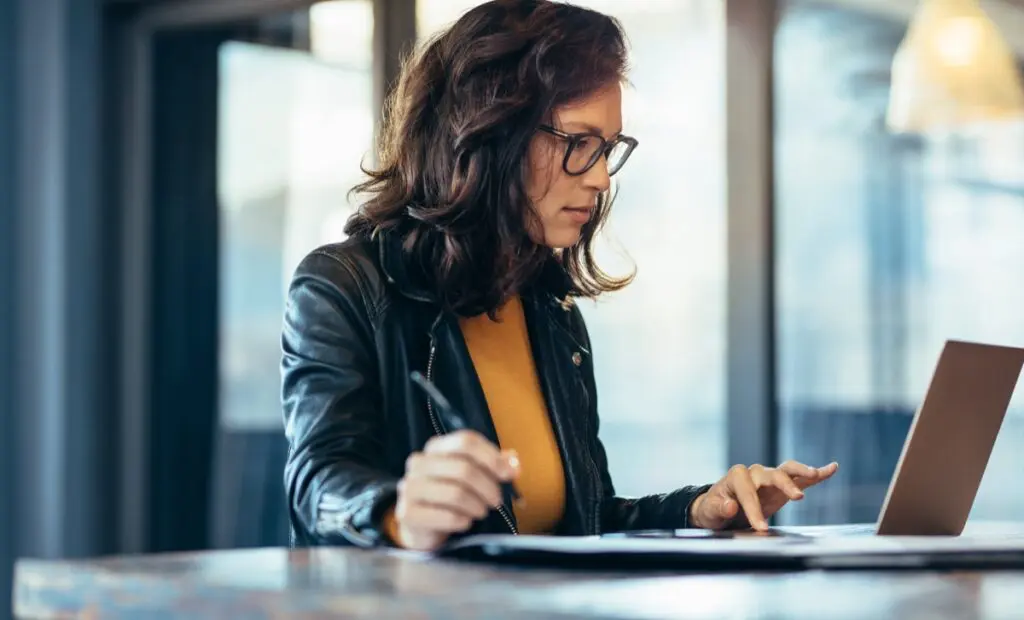 Businesswoman making notes looking at a laptop computer at office. Woman entrepreneur sitting at the table writing notes while working on laptop.